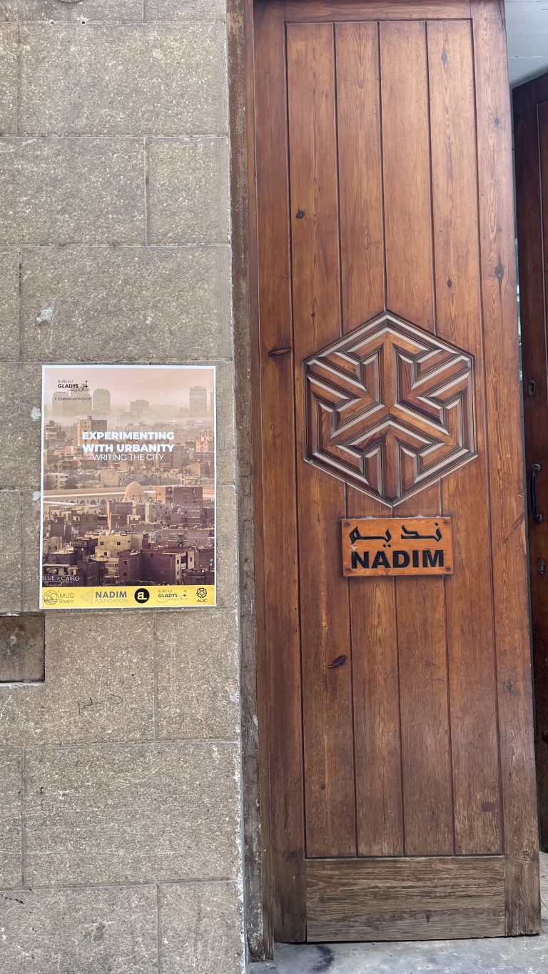 A carved wooden door with the Experimenting with Urbanity workshop poster on the stone wall beside it