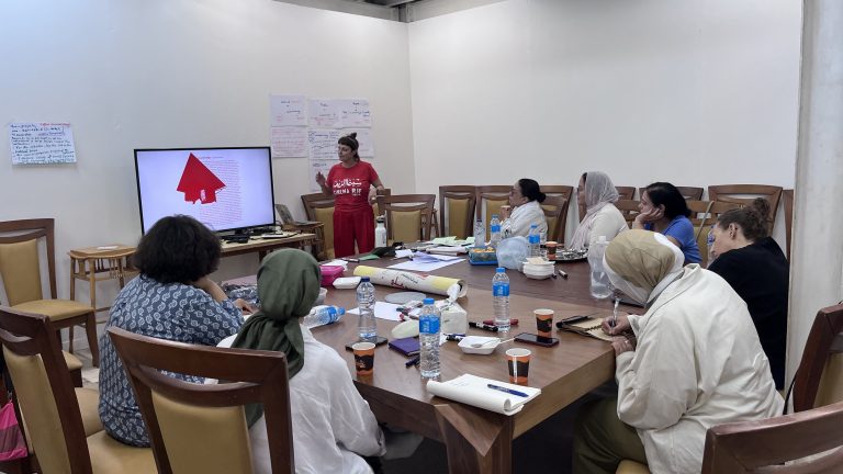 A group of women sit around a large wooden conference table during a workshop session. One woman in a red shirt stands at the head of the table, presenting a red graphic on a television screen while others take notes. Several water bottles, coffee cups, and papers are scattered across the table.