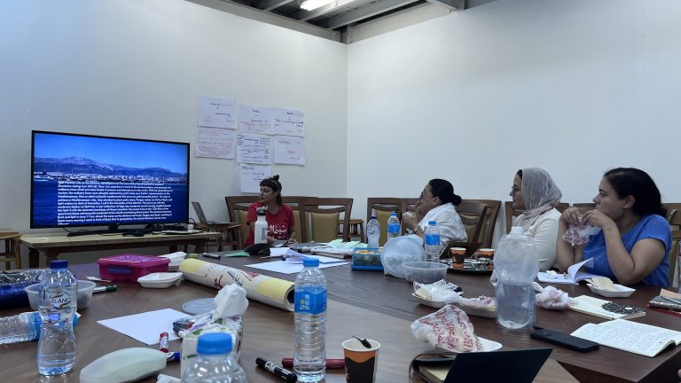 A close-up view of participants seated at a conference table during a seminar. A television in the background displays a coastal cityscape with detailed text overlay. The table is cluttered with notebooks, water bottles, and snack wrappers.