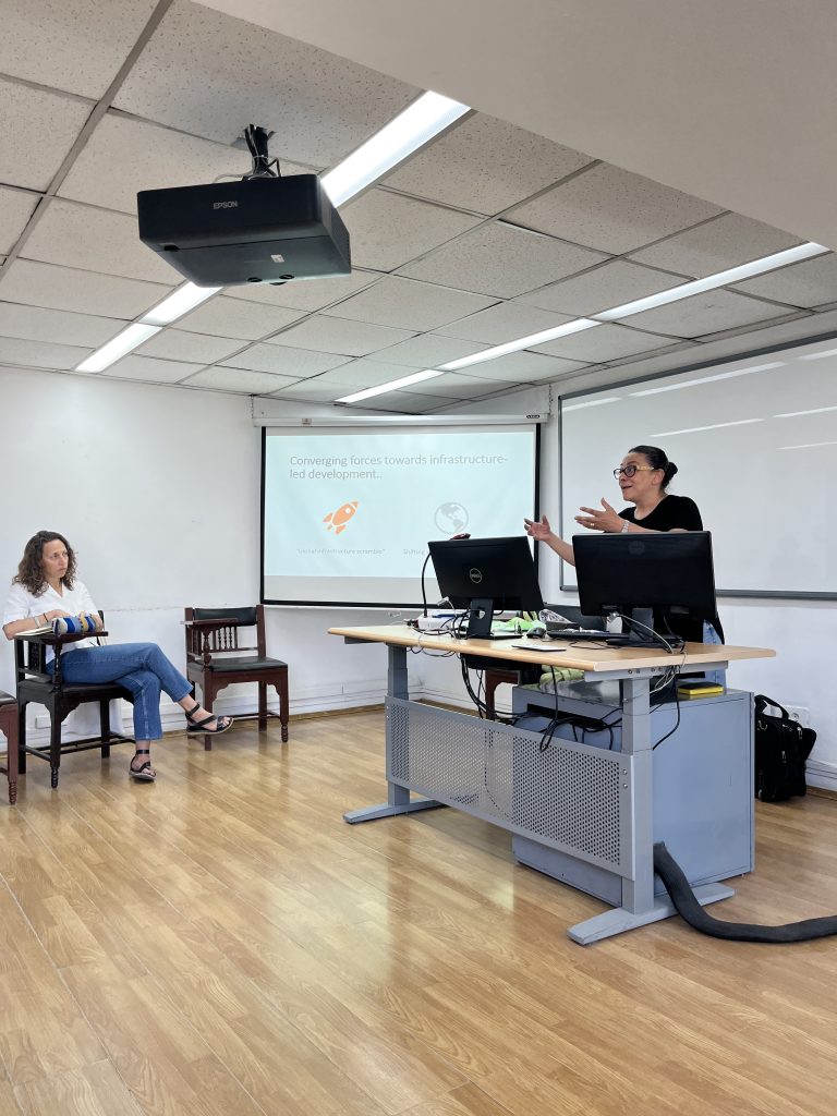 A speaker at a lectern beside a projected slide during the Experimenting with Urbanity workshop