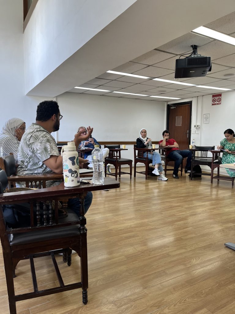 Several participants are seated in a circle in a bright room with wooden floors, engaging in a group discussion. One man in a patterned shirt is gesturing while speaking. Each person sits in a dark wooden chair with an attached writing desk.