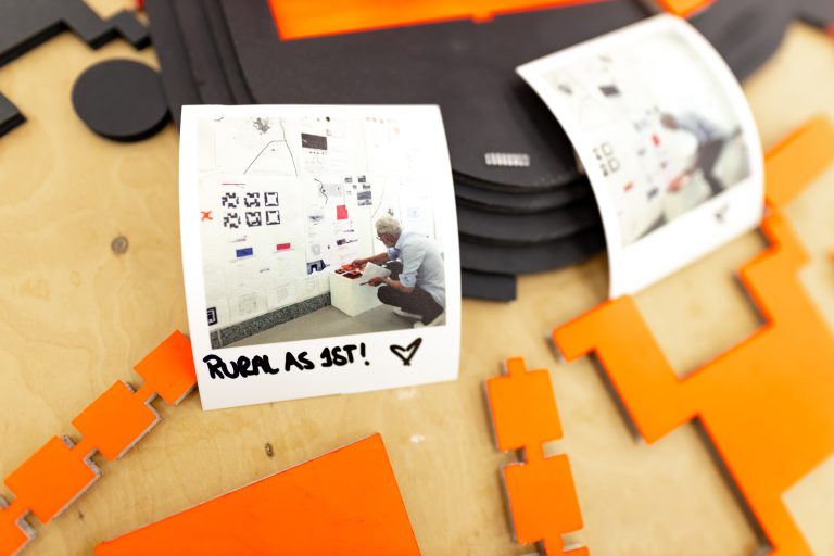 Printed photo cards on a wooden table with orange cut-out shapes at the Among Us exhibition